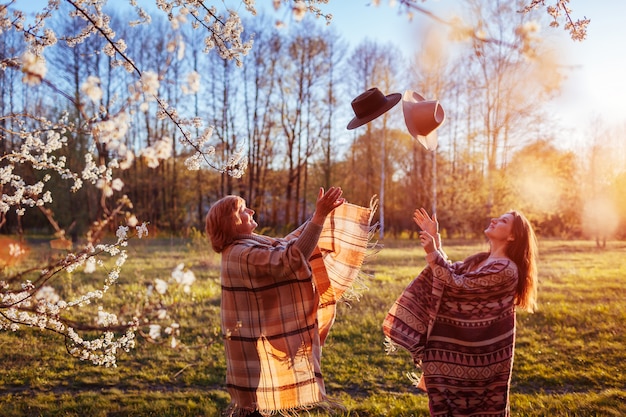 Madre de mediana edad y su hija adulta arrojando sombreros en jardín floreciente. Mujeres divirtiéndose