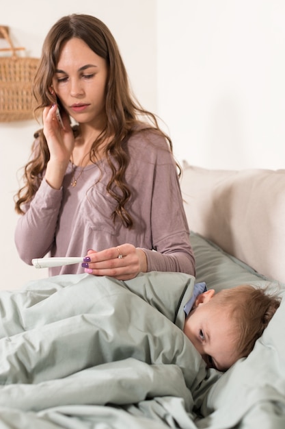 Madre midiendo la temperatura de su hijo y llamando a un médico. Niño enfermo se siente mal y acostado en la cama debajo de una manta en casa. Coronavirus pandémico, covid-19.