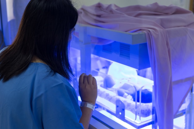 Madre mirando al bebé recién nacido con hiperbilirrubinemia / ictericia neonatal bajo luz ultravioleta azul