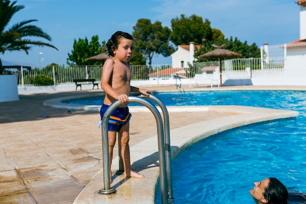 Madre mirando a su hijo desde el interior de la piscina, mientras el niño está afuera aferrado a la escalera mojada