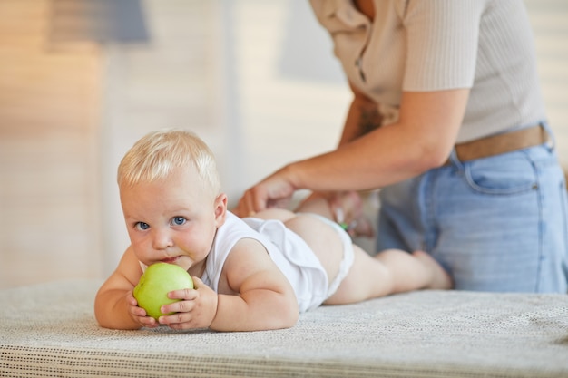 Madre moderna cambiando de ropa de su pequeño hijo mientras él muerde la manzana verde