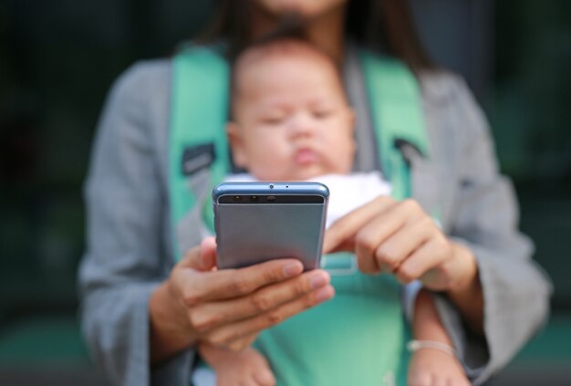 La madre del negocio en juego está utilizando smartphone con llevar a su bebé en hipseat.