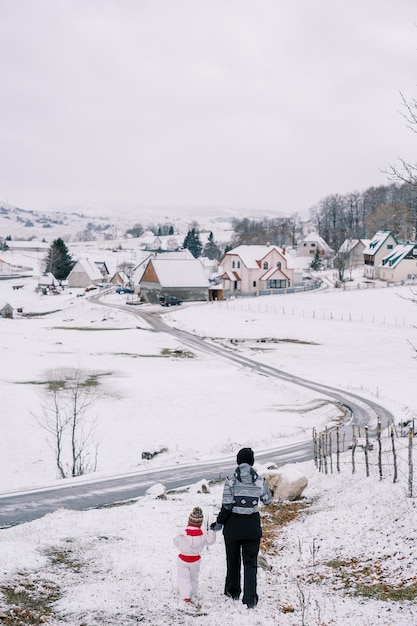 Madre y una niña caminan por una colina nevada tomados de la mano hacia una vista trasera de la carretera del pueblo