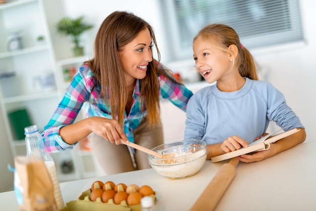 La madre y la niña mezclan masa en la cocina doméstica y la hija está leyendo la receta del libro de cocina. La madre aprende a su hija a hacer las galletas.