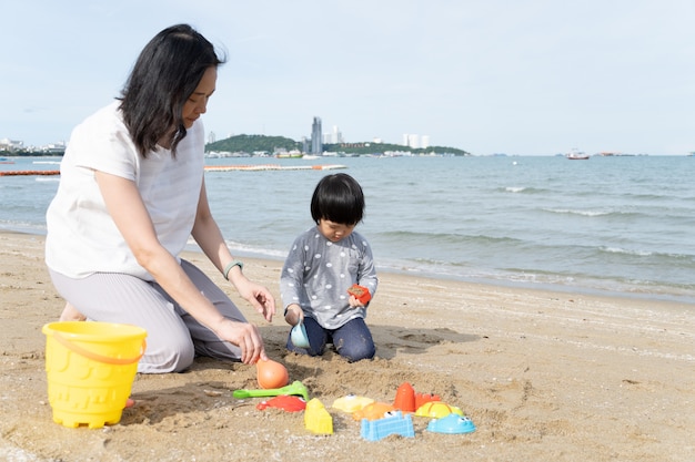 Madre y niño asiático de 3 años juegan arena en la playa.