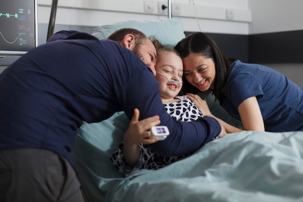 Foto madre y padre alegres abrazando a una hija enferma sentada en la cama de un paciente mientras está bajo tratamiento. niña enferma feliz abrazada por padres alegres y sonrientes en la sala de pediatría del hospital.