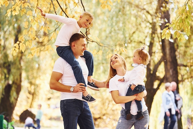 La madre y el padre sostienen a los niños en los hombros y en las manos. Familia joven alegre dar un paseo juntos en un parque de otoño.
