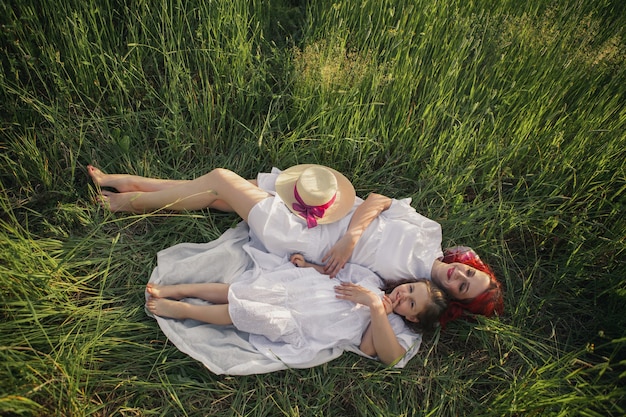 Madre con el pelo rojo y un sombrero se encuentra en un campo verde con césped con su hija de cuatro años al atardecer