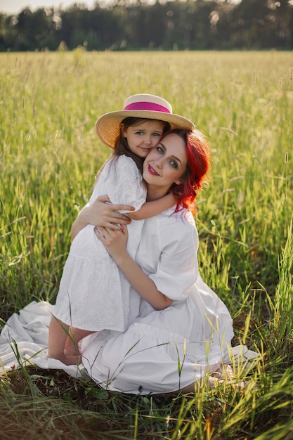 Foto madre con el pelo rojo y un sombrero está sentada en un campo verde con césped con su hija de cuatro años al atardecer