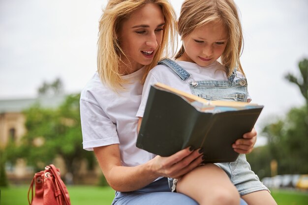 Madre pensativa pasar tiempo con su hija en el interesante libro mientras está sentada en el parque de la ciudad