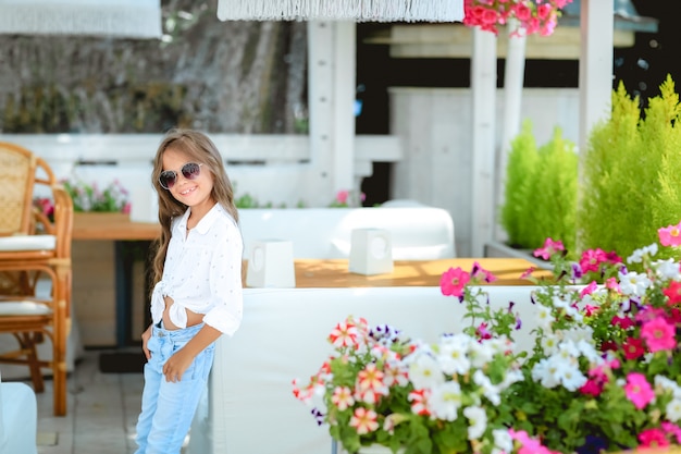 Madre con pequeña hija encantadora están sentados en la terraza de verano en la luz del sol. Ellos tienen brunch. Centrarse en la niña. Emociones reales La niña está mirando a la cámara. Hermoso retrato