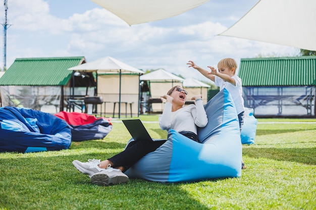 Foto madre con portátil, su hijo jugando al aire libre en el césped verde descanse y trabaje con un niño en el parque