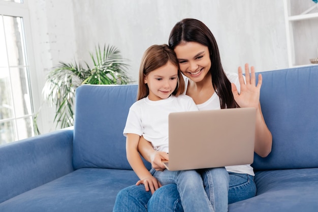 Madre positiva usando una computadora portátil y agitando su mano durante una conversación de video mientras mantiene a su pequeña hija de rodillas