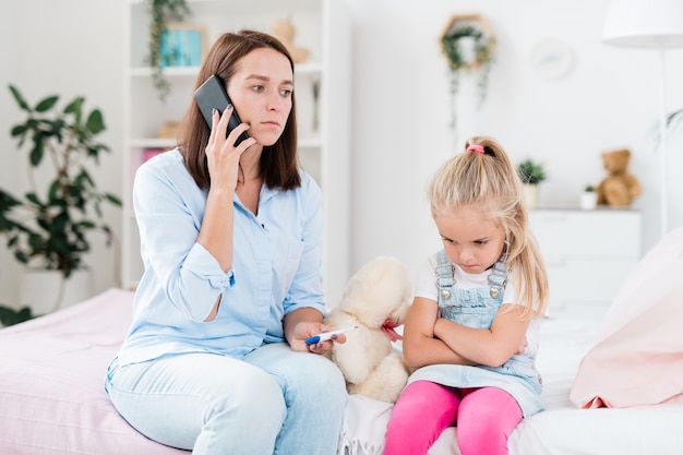 Madre preocupada con termómetro y teléfono inteligente llamando al médico a casa mientras está sentada junto a su pequeña hija enferma