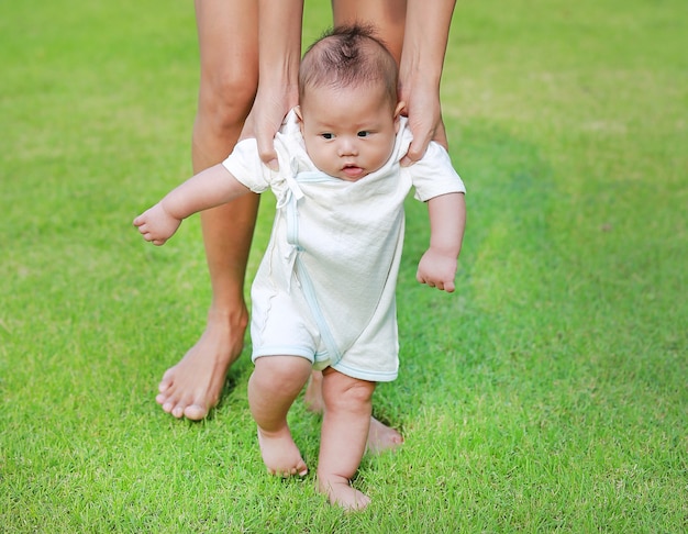 Madre que entrena a su bebé infantil para caminar primeros pasos en el jardín de la hierba verde.