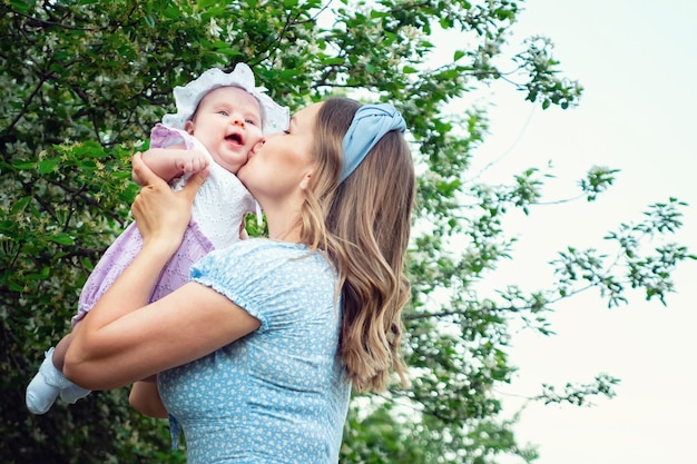 Madre rubia alegre lanza una niña divertida y besa a un niño de pie en el parque local contra árboles verdes con flores blancas en primavera, copyspace