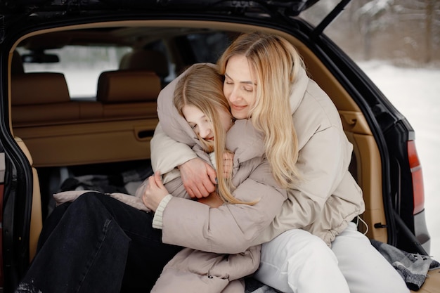 Madre rubia e hija adolescente sentadas en el maletero del coche abierto en el bosque de invierno