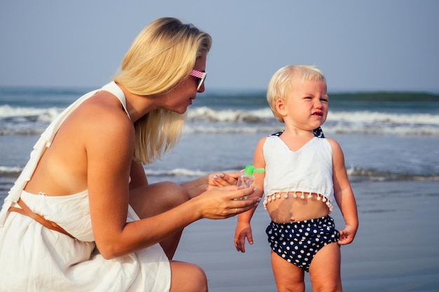 La madre rubia con un vestido blanco y su hija rubia de un año usan un gel antiséptico antibacteriano en la playa del océano Índico