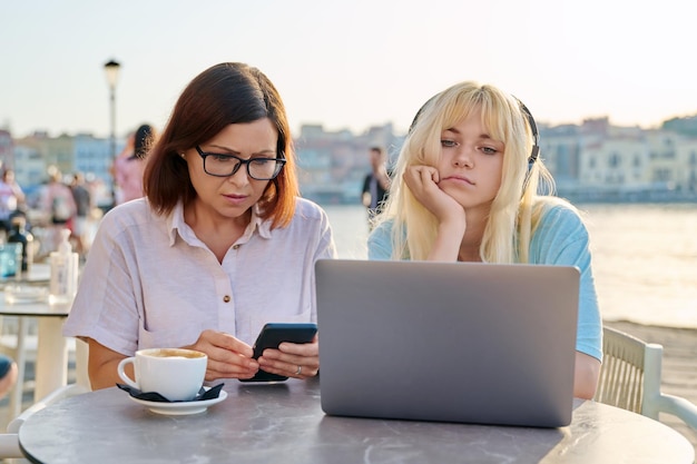 Madre seria e hija adolescente juntas en un café al aire libre mirando una laptop