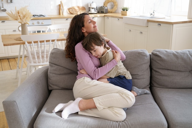 La madre solícita y el niño preescolar se sientan en el sofá abrazándose con los ojos cerrados, amor familiar y unión