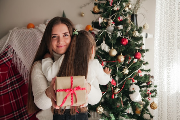 Madre sonriente abrazando a su hija con regalos en sus manos y adornos navideños en el fondo concepto de regalos familiares navideños