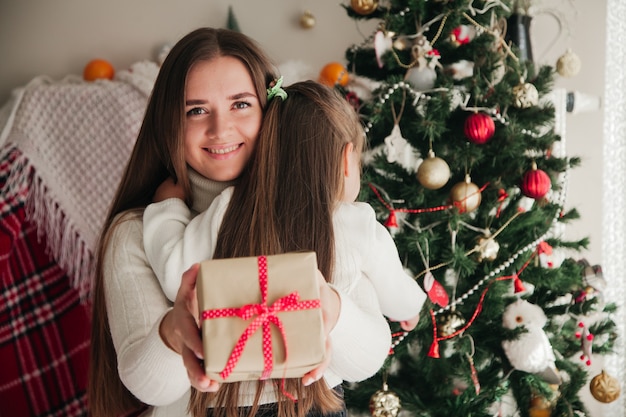 Madre sonriente abrazando a su hija con regalos en sus manos y adornos navideños en el fondo concepto de regalos familiares navideños