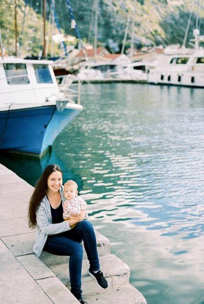 Madre sonriente con un bebé en su regazo se sienta en el muelle cerca del barco