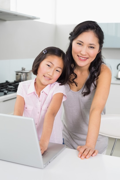 Madre sonriente con hija joven que usa la computadora portátil en cocina