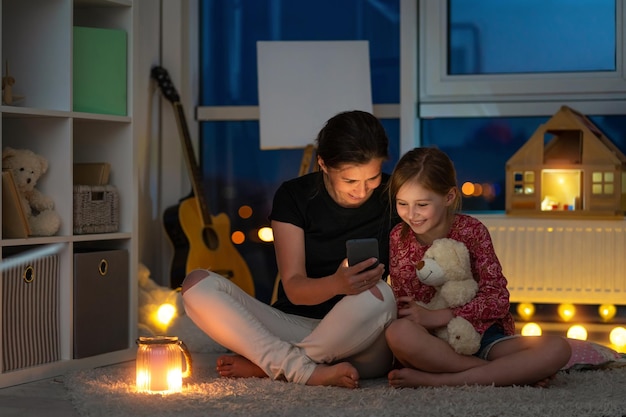 Madre sonriente con una hija pequeña usando un teléfono inteligente sentado en el suelo en una habitación nocturna iluminada