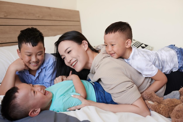 Madre sonriente y sus tres hijos jugando en la cama y haciéndose cosquillas y riendo