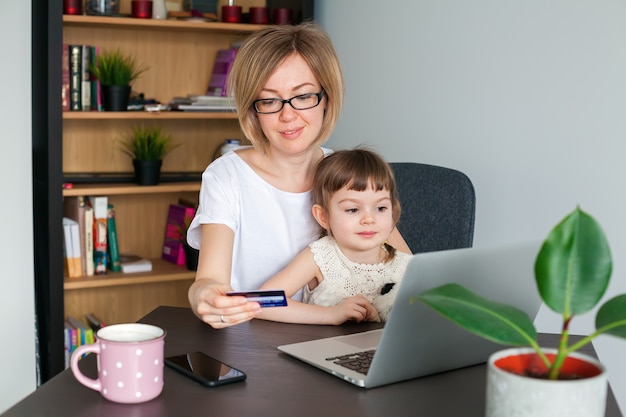 Madre sosteniendo una tarjeta de crédito con su pequeña hija sentada cerca de mirar la computadora portátil. Concepto de compras en línea.