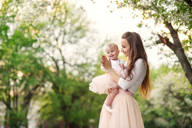 La madre sostiene a su pequeña hija en sus brazos entre los árboles en flor. Mamá y su pequeño bebé lucieron un vestido rosa de aspecto familiar.