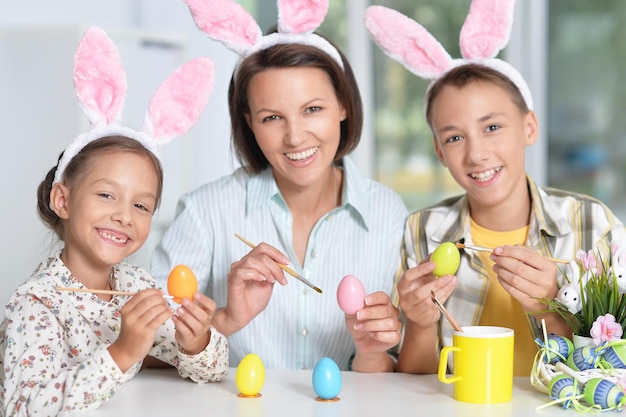 Madre con su hijo e hija vistiendo orejas de conejo pintando huevos de Pascua