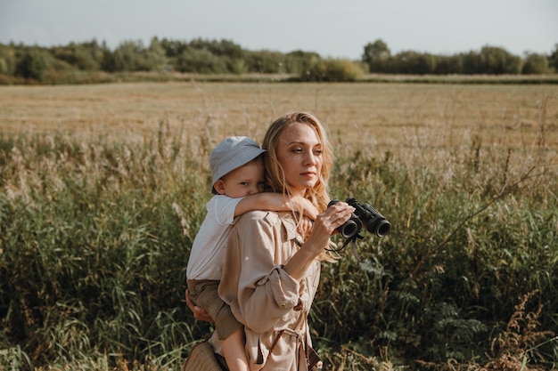 Una madre con su hijo en la espalda está caminando en la naturaleza sosteniendo binoculares en sus manos