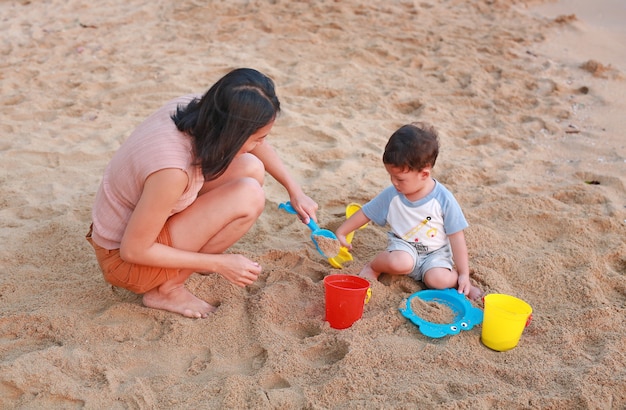 Madre y su hijo jugando arena en la playa con juguete de plástico