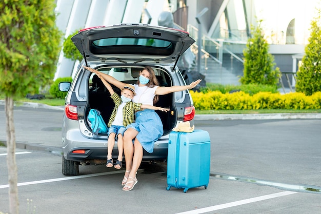 Una madre y su hijo con máscaras médicas y una maleta azul se van de vacaciones o viajan sentados en un camión frente al aeropuerto con las manos extendidas como un avión