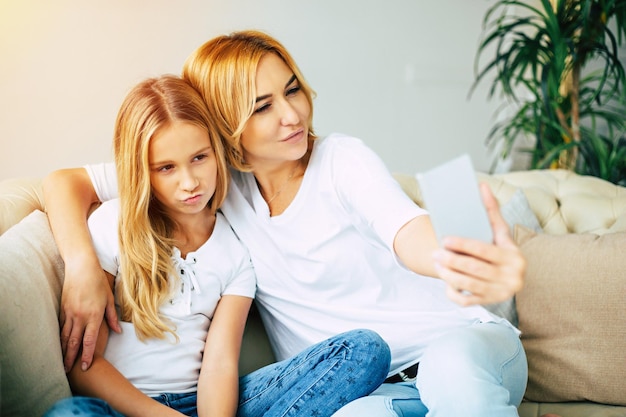 La madre y su pequeña hija se abrazan, posan y hacen una foto selfie en un teléfono inteligente mientras se sientan en el sofá de casa