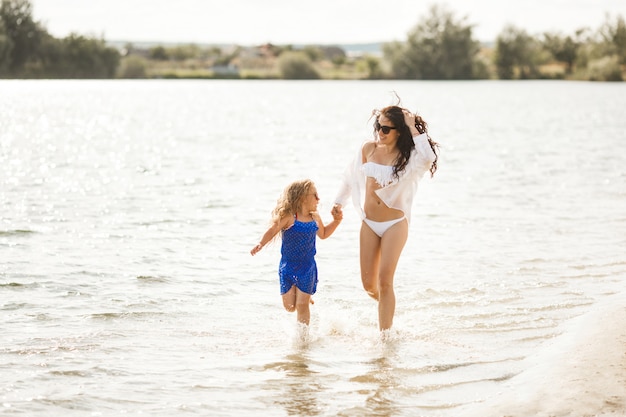 Madre y su pequeña hija divirtiéndose en la costa. Mamá bonita joven y su hijo jugando cerca del agua