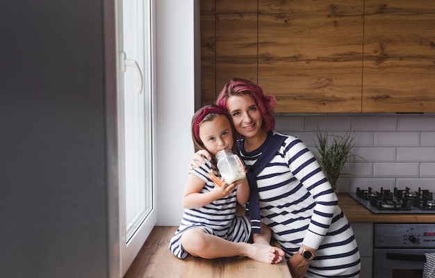 madre y su pequeña hija están sentadas en el alféizar de la ventana de la cocina y beben leche