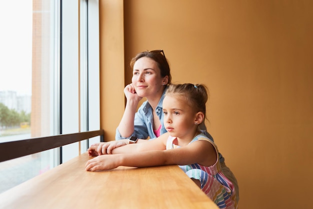 Madre con su pequeña hija sentada cerca de la ventana del café. esperando la orden