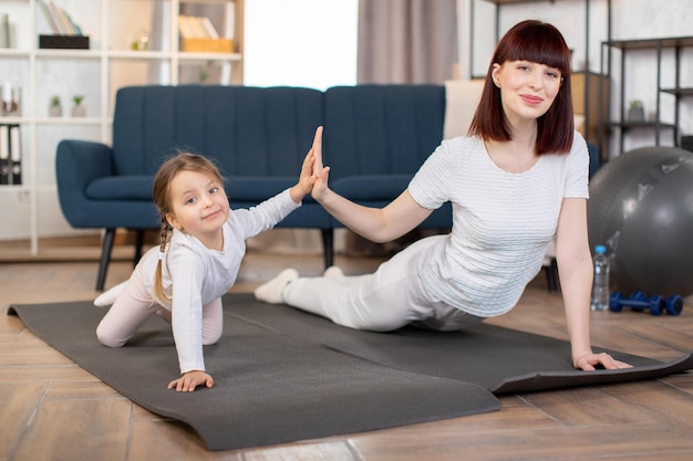 La madre y su pequeña y linda hija hacen ejercicios de flexiones en la alfombra de yoga en la sala de estar en casa
