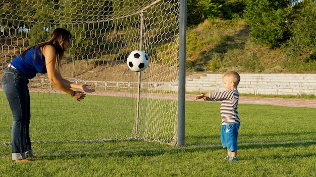 Madre y su pequeño hijo de pie delante de los postes de la portería en un campo de deportes de hierba verde lanzando una pelota de fútbol entre sí