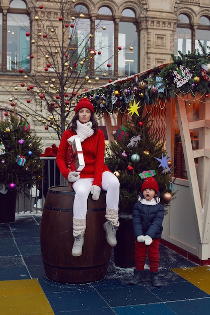 Foto madre en un suéter de punto rojo y sombrero con un regalo de pie al lado de su hijo gracioso en la calle al lado del árbol de navidad