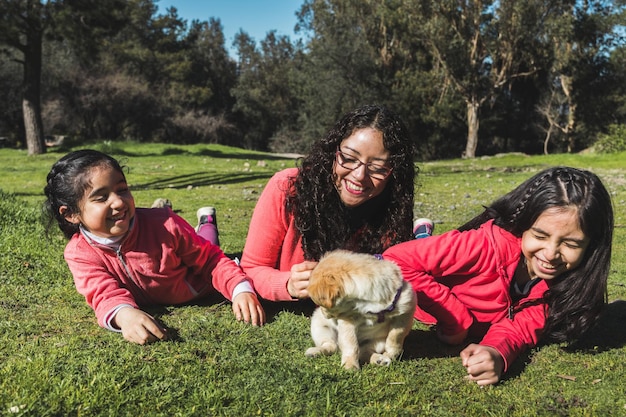 Madre con sus dos hijas tiradas en el césped y jugando con un cachorro golden retriever en el parque