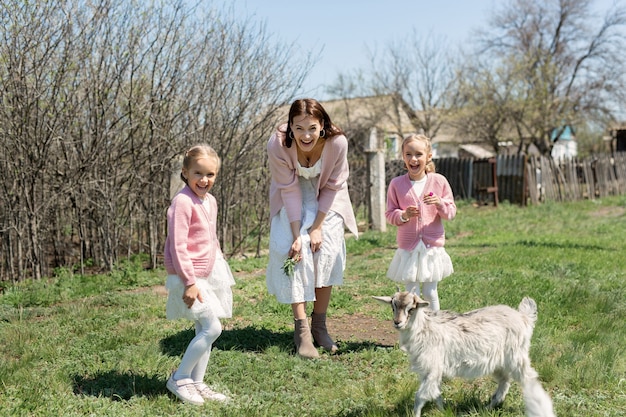 Madre con sus hijas gemelas alimenta a una cabra en un prado de la aldea.