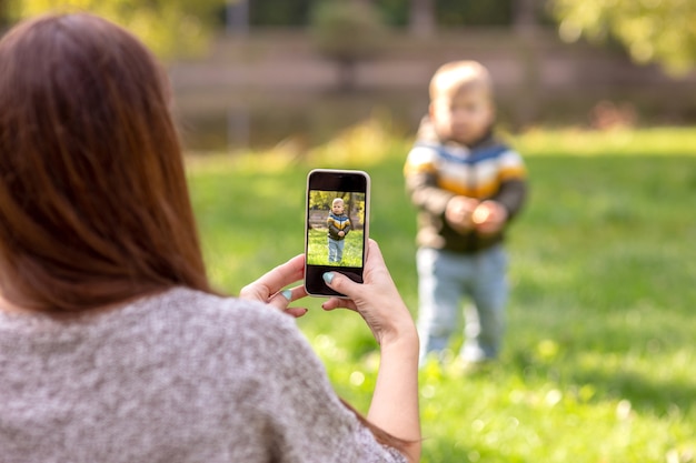 Una madre toma fotografías de su pequeño hijo en un teléfono inteligente en un parque verde