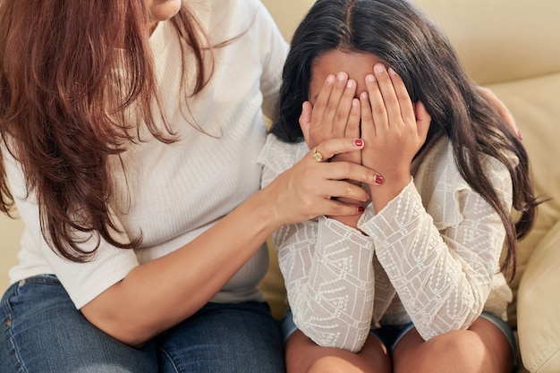 Madre tratando de consolar a su hija cubriéndose la cara con las manos y llorando después de ser intimidada en la escuela