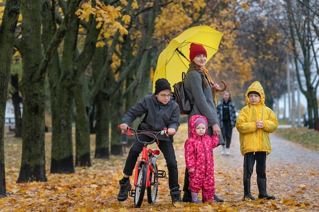 Madre con tres hijos de pie en el parque en el parque de otoño. Callejón con hojas caídas.