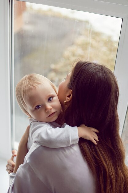 Madre con una túnica blanca se sienta con un niño una hija rubia en una ventana grande de la casa