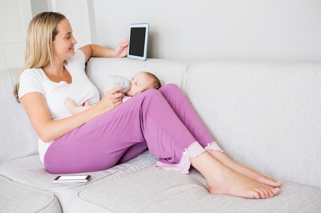 Madre usando tableta digital mientras alimenta a su bebé con biberón